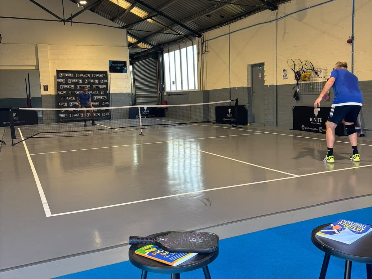 A daytime pickleball rally on the grey court at Ignite, with rackets hung on the wall behind