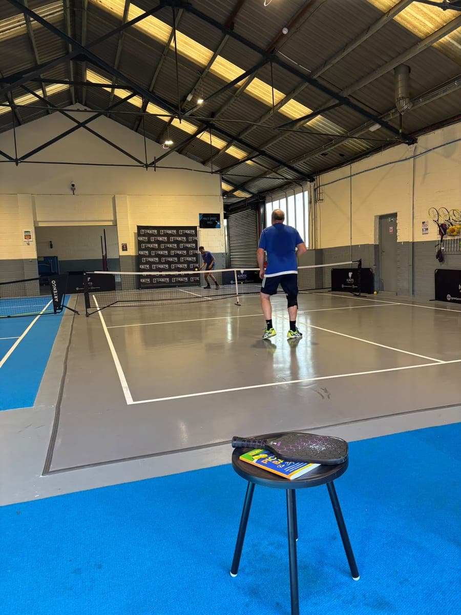 A daytime pickleball rally at Ignite, seen from the lounge, with a paddle and a beginner's guide to pickleball on a stool in front
